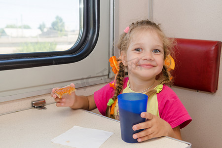 一张笑脸的小女孩在火车外二等车厢的桌子旁喝茶和三明治