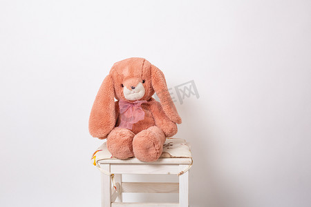 兔子粉色摄影照片_粉红色的兔子毛绒娃娃坐在白色背景的凳子上。