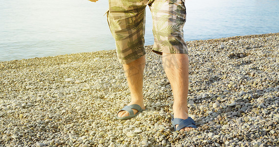 男人的脚穿着人字拖和短裤，一个男人站在卵石滩上的沙滩上，早上日出