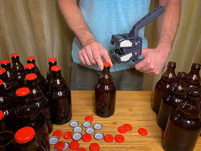 啤酒瓶啤酒盖摄影照片_在家里酿造精酿啤酒，男人在木桌上用塑料封盖器关闭棕色玻璃啤酒瓶，上面有红色皇冠盖。