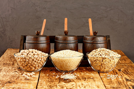 鹰嘴豆、米饭和燕麦片放在玻璃碗里，背景是小木桶，用于在棕色背景下的旧木桌上储存散装产品。