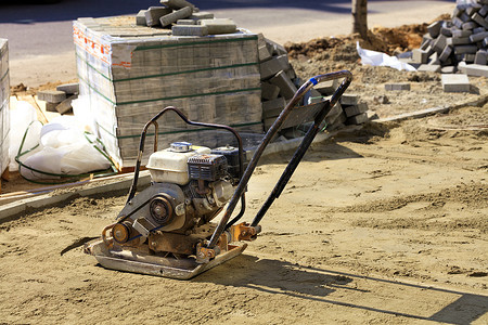 一堆铺路石对面是一台用于压实沙土的旧汽油压实机。