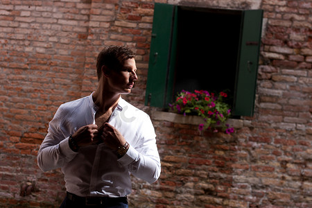 带窗户的砖墙摄影照片_威尼斯身穿白衬衫的男子靠着砖墙和一扇带鲜花的美丽窗户 砖墙背景下的威尼斯模特
