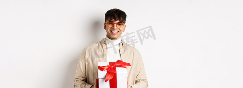 英俊的年轻人制作礼物，拿着装有礼物的盒子，对着镜头微笑，站在白色背景上