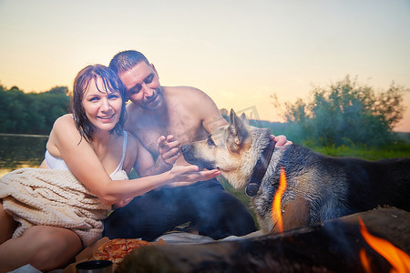 快乐的湿夫妇放松，在夏日阳光明媚的傍晚在日落时在大自然中露营，在火边与大狗玩耍和拥抱。