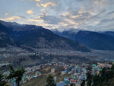 季风云在积雪覆盖的喜马拉雅山脉上移动，蓝橙色日落日出光与山脚下的库鲁马纳利山谷镇