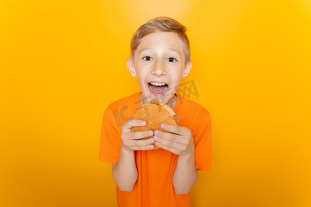 吃汉堡的男孩摄影照片_一个穿着橙色 T 恤的男孩面前拿着一个汉堡，在黄色背景下大声笑