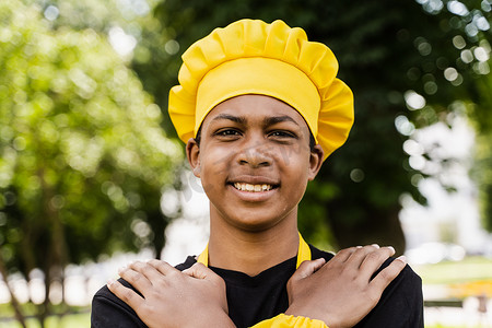 非洲黑人青少年戴着厨师帽，穿着黄色围裙制服，在户外微笑。