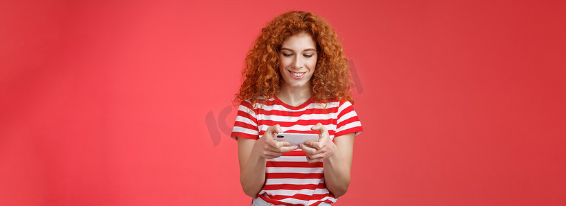 红发女孩在等待队列牙医周围玩耍，玩很棒的智能手机游戏，握住电话水平点击手机屏幕，看起来电话显示屏微笑高兴娱乐站立红色背景