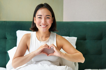平静的心摄影照片_微笑的韩国女孩表现出心、爱的标志，花时间在卧室里，早上躺在床上，盖着白色羽绒被
