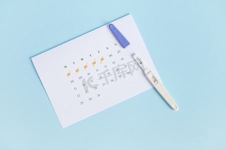 顶视图阳性妊娠试验和白色日历，最后几天的月经被标记，隔离在蓝色背景上。