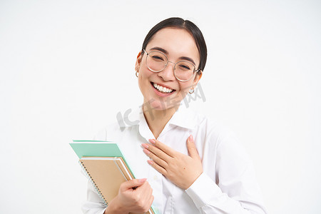 放在胸前摄影照片_戴着眼镜的友善快乐的亚洲女性，老师把手放在胸前微笑，自我介绍，站在白色背景中