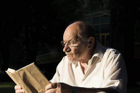一位身穿白衬衫的老人坐在公园的地上的毯子上，正在读一本有趣的书。