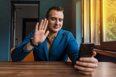 一位年轻时尚的男士，欧洲外貌肖像的商人，穿着西装坐在桌旁，通过智能手机的视频通信进行交谈，用手势发送问候