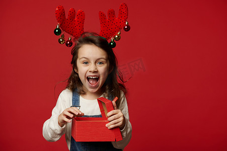 带着鹿角圈的喜出望外的顽皮女婴对着镜头大笑，在红色背景下摆出圣诞礼物的姿势
