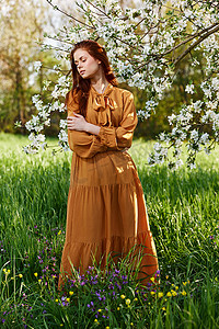 一位苗条、甜美的女人穿着橙色长裙，站在一棵开花的树附近的高草丛中，双臂交叉在胸前，目光移向别处