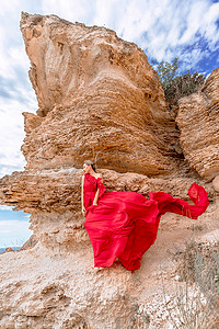 丝绸背摄影照片_一位身着红色丝绸连衣裙的女子站在海边，背靠群山，裙子在微风中摇曳。