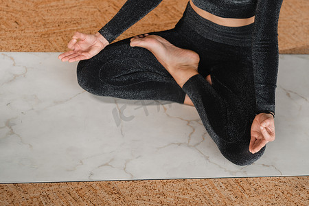 一位身穿黑色运动服的女性坐在健身房的莲花坐瑜伽垫上的腿和手臂的特写