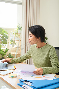 集中精力的亚洲女性坐在桌子旁，在家庭办公室的笔记本电脑上做笔记