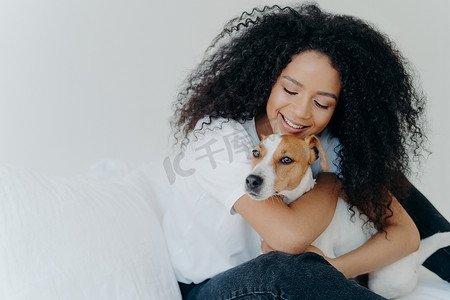 一起小狗在睡觉摄影照片_快乐的非洲裔美国女人表达对狗的爱，拥抱宠物，穿着休闲服，坐在卧室舒适的床上，与家畜一起睡觉。