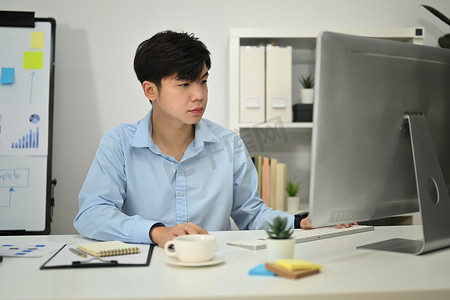 集中精力摄影照片_亚洲男性投资者集中精力，看着电脑显示器分析办公室的财务数据