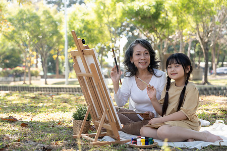 快乐可爱的亚洲祖母教她的孙女在画布上画水彩，在美丽的公园里度过了愉快的时光
