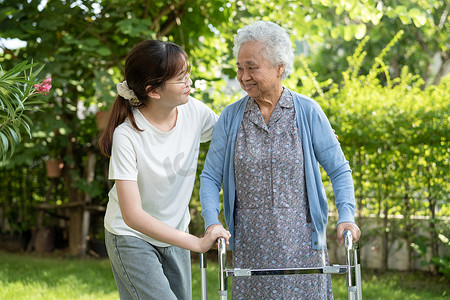 护理人员帮助亚洲老年女性患者在公园里用步行器行走，健康强大的医疗理念。