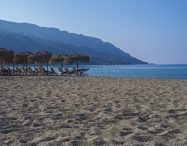 太阳伞摄影照片_希腊科孚岛 Agios Georgios Pagon 的沙子和卵石海滩和绿松石海岸，配有太阳伞橙色日光浴床和船、绿色山丘和清澈的蓝天背景