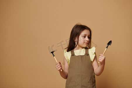 可爱的小女孩手里拿着花园铲和耙子，孤立的米色背景。