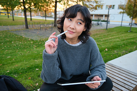 时尚艺术字摄影照片_亚洲女性的肖像，公园里的学生，坐在长凳上，拿着数字平板电脑和笔，思考，一边思考，一边做笔记，在户外做作业