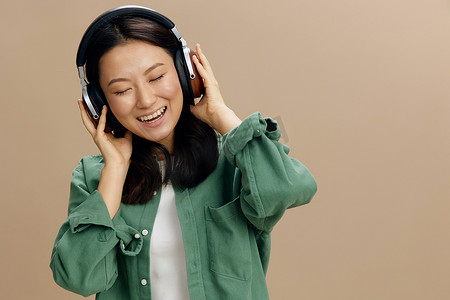 放松地享受最喜欢的歌曲可爱的亚洲学生年轻女子穿着卡其绿色衬衫触摸耳机在米色柔和的工作室背景上摆出孤立的姿势。
