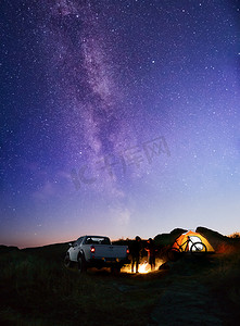 朋友们靠近篝火、皮卡车、帐篷和自行车在夜空下的山上与银河。
