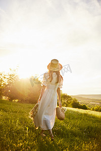 一位身穿浅色长裙的女子头戴帽子，手里提着篮子，在夕阳的照射下漫步在乡村，享受着大自然。