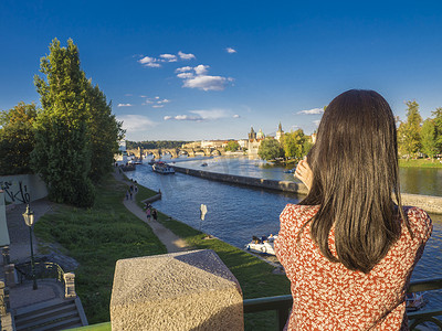 年轻女子，用手机从军团桥拍照，欣赏坎帕岛、伏尔塔瓦河、布拉格城堡、老城区、夏日阳光明媚的午后、捷克共和国、布拉格、夏日阳光明媚的日子
