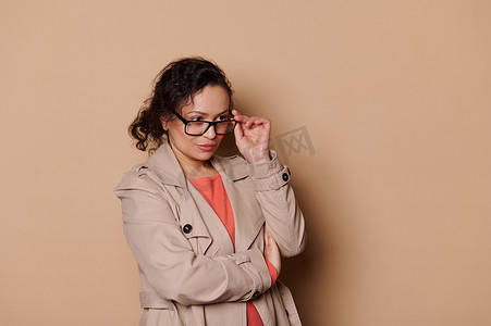 眼科广告摄影照片_米色背景上的孤立肖像，是一位好奇的黑发中年女士，她戴着眼镜，看着广告空间