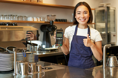 微笑的亚洲女孩咖啡师，围裙的咖啡馆老板，展示刷卡机，支付读卡器，在咖啡店接受非接触式订单
