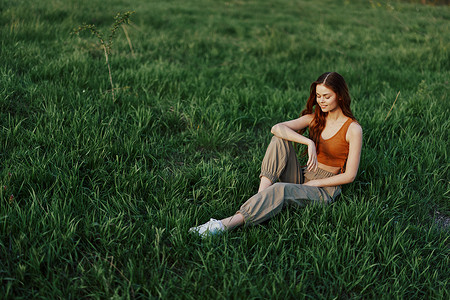 红发女人坐在公园的绿草地上，穿着橙色上衣、绿色裤子和运动鞋，看着夏日的夕阳