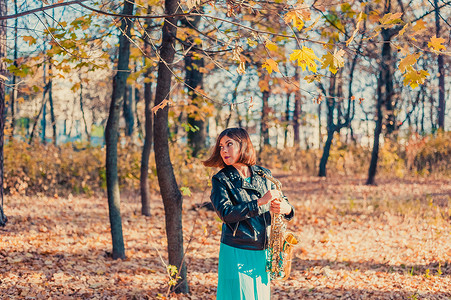 黄色秋季公园里，穿着黑色夹克和蓝色裙子、拿着萨克斯管的漂亮黑发女孩