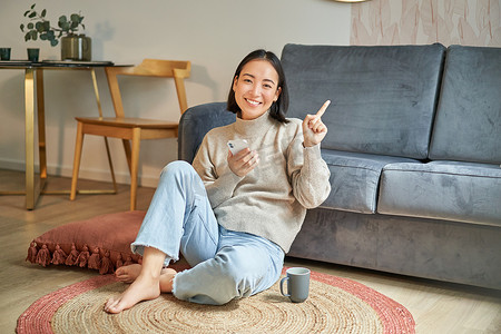 促销房屋摄影照片_微笑的亚洲女孩坐在时尚客厅的地板上，指着广告，展示促销横幅，手里拿着手机