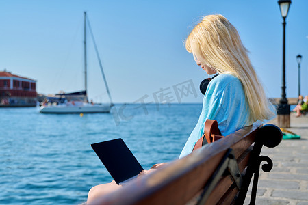 海边女子摄影照片_在海边戴着笔记本电脑、戴着耳机放松的年轻女子