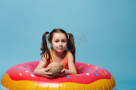 美丽的小女孩，拿着椰子鸡尾酒，在游泳圈甜甜圈里，在孤立的蓝色背景下对着镜头微笑。