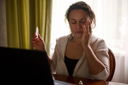 药房摄影照片_多民族妇女坐在桌旁，拿着笔记本电脑，使用药用眼药水缓解干眼和数码眼疲劳