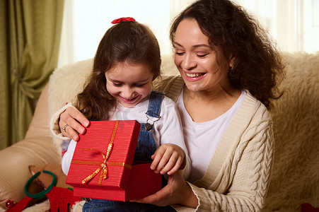 小女孩，可爱的女儿坐在妈妈的膝盖上，在打开圣诞礼盒时体验快乐和兴奋