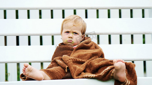 悲伤的贫穷小女孩摄影照片_夏天，一个一岁的小女孩，裹着毛巾，脏兮兮的脸，孤独地坐在花园的秋千上。