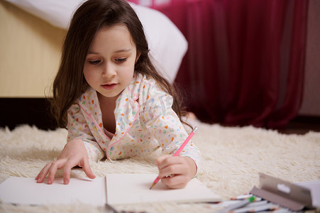 穿着睡衣的特写小女孩，躺在卧室的地毯上，专注于用彩色铅笔画画