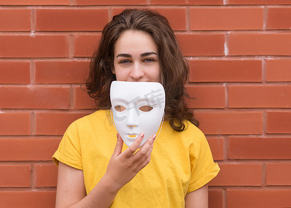 戴情绪面具的人摄影照片_身穿黄色衬衫的年轻女子在砖墙上戴上面具