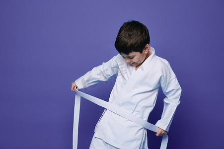 儿童跆拳道摄影照片_自信快乐的小男孩将合气道和服的白色腰带系在腰上。