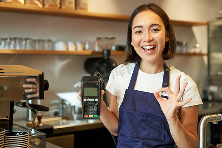 微笑的亚洲咖啡师女孩，穿围裙，展示信用卡机处理付款，建议非接触式付款，站在咖啡店