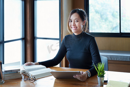 一名亚洲少女使用平板电脑和书籍通过图书馆木桌上的视频会议在线学习的肖像