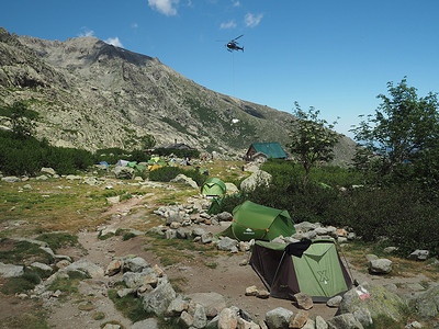 法国，科西嘉岛，科西嘉阿尔卑斯山，2017年6月19日：直升机在著名的远足小径GR20、岩石、帐篷、树木和蓝天背景上为山地营地Refuge de Pietra Piana运送物资
